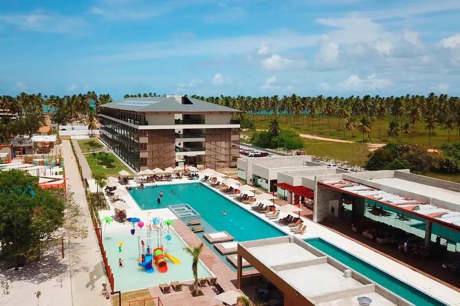 An aerial view captures a modern resort surrounded by lush greenery and palm trees. A long swimming pool is visible, lined with sun loungers and umbrellas. Colorful water play equipment is present, adding vibrancy to the pool area, with sandy beach and ocean in the background.