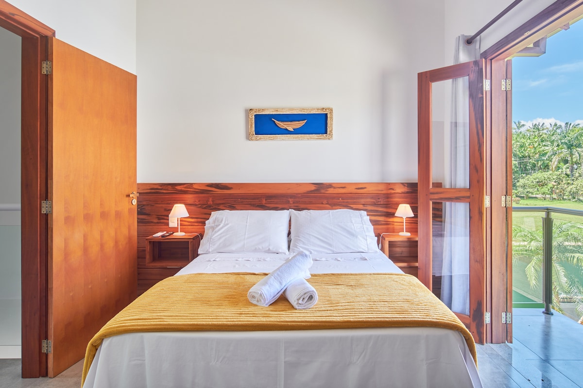 A well-lit bedroom features a spacious bed with fresh linens and neatly arranged towels. Natural light floods the room through open double doors, revealing views of greenery outside. The modern wooden wall and simple artwork create a calm atmosphere.