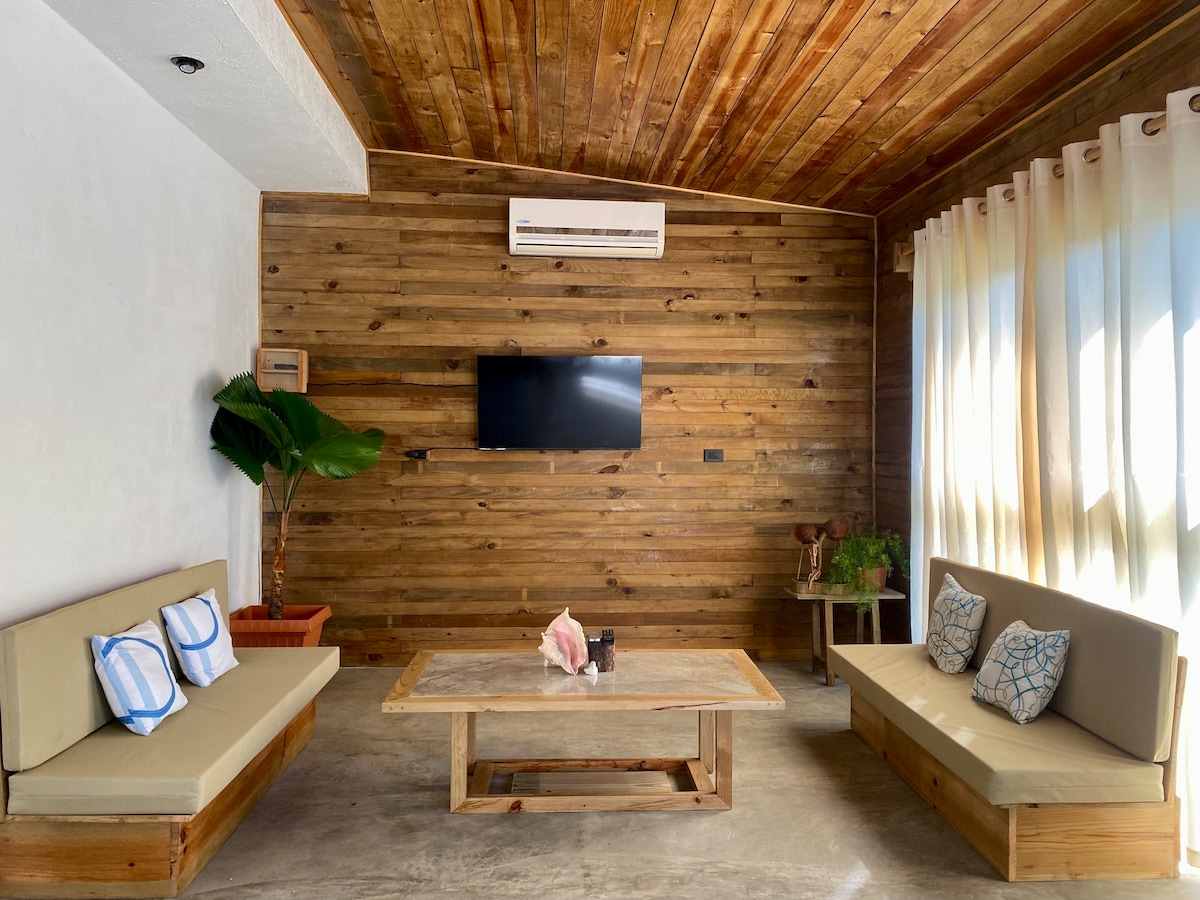 A welcoming living area features wooden walls and a ceiling, creating a natural ambiance. Two comfortable benches are positioned around a rustic coffee table. A flat-screen television is mounted on the wall, while large windows allow natural light to fill the space.