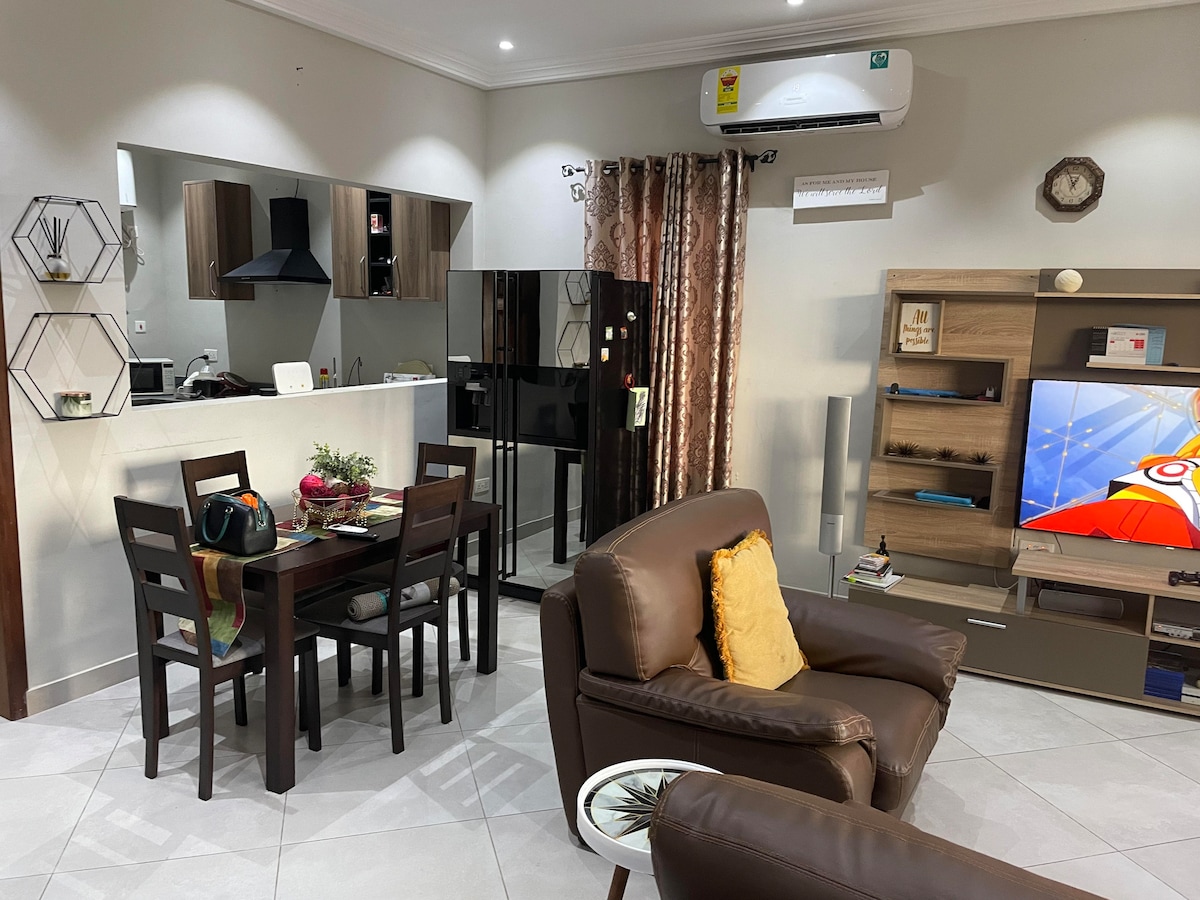 An open living area is featured, showcasing a dining table set for four adjacent to a kitchen with modern appliances. A comfortable seating arrangement is positioned in the foreground, and a television stand is visible against the wall, providing a cozy atmosphere.