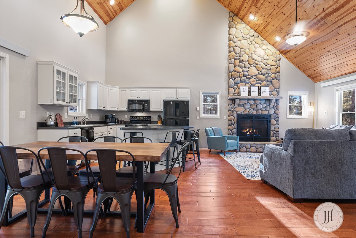 An open-concept living area features a high, vaulted ceiling with wooden beams. A stone fireplace is the focal point, complemented by a cozy seating arrangement. A large dining table with metal chairs is visible, along with a fully equipped kitchen, creating a warm, inviting space.