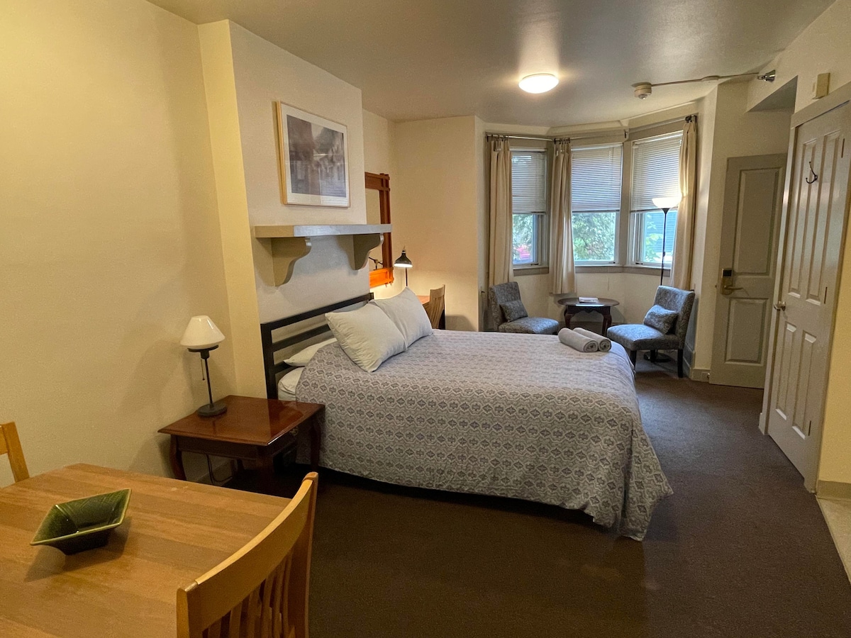 A spacious studio room is presented, featuring a queen-sized bed with a patterned blue coverlet. Natural light enters through two windows beside a pair of comfortable chairs. A small wooden dining table is positioned next to the bed, enhancing the room's functionality.