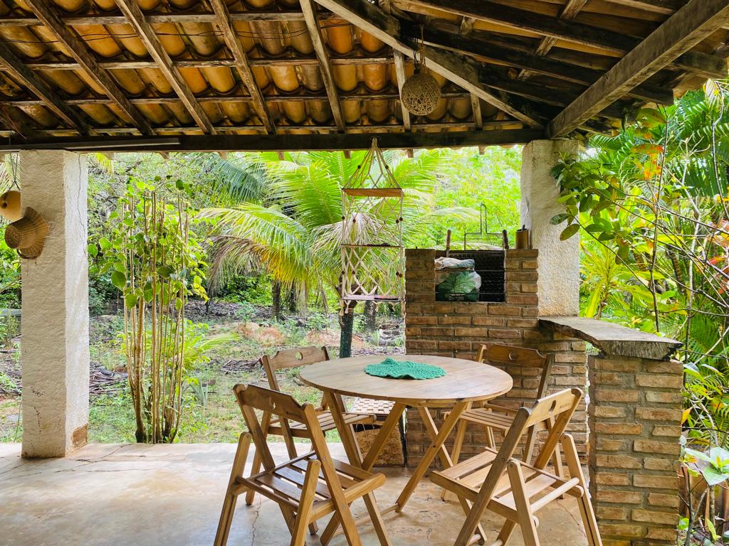An outdoor seating area is nestled under a rustic roof, featuring a round wooden table surrounded by four folding chairs. A brick barbecue grill is positioned nearby, with tropical greenery in the background, creating a natural and inviting setting.