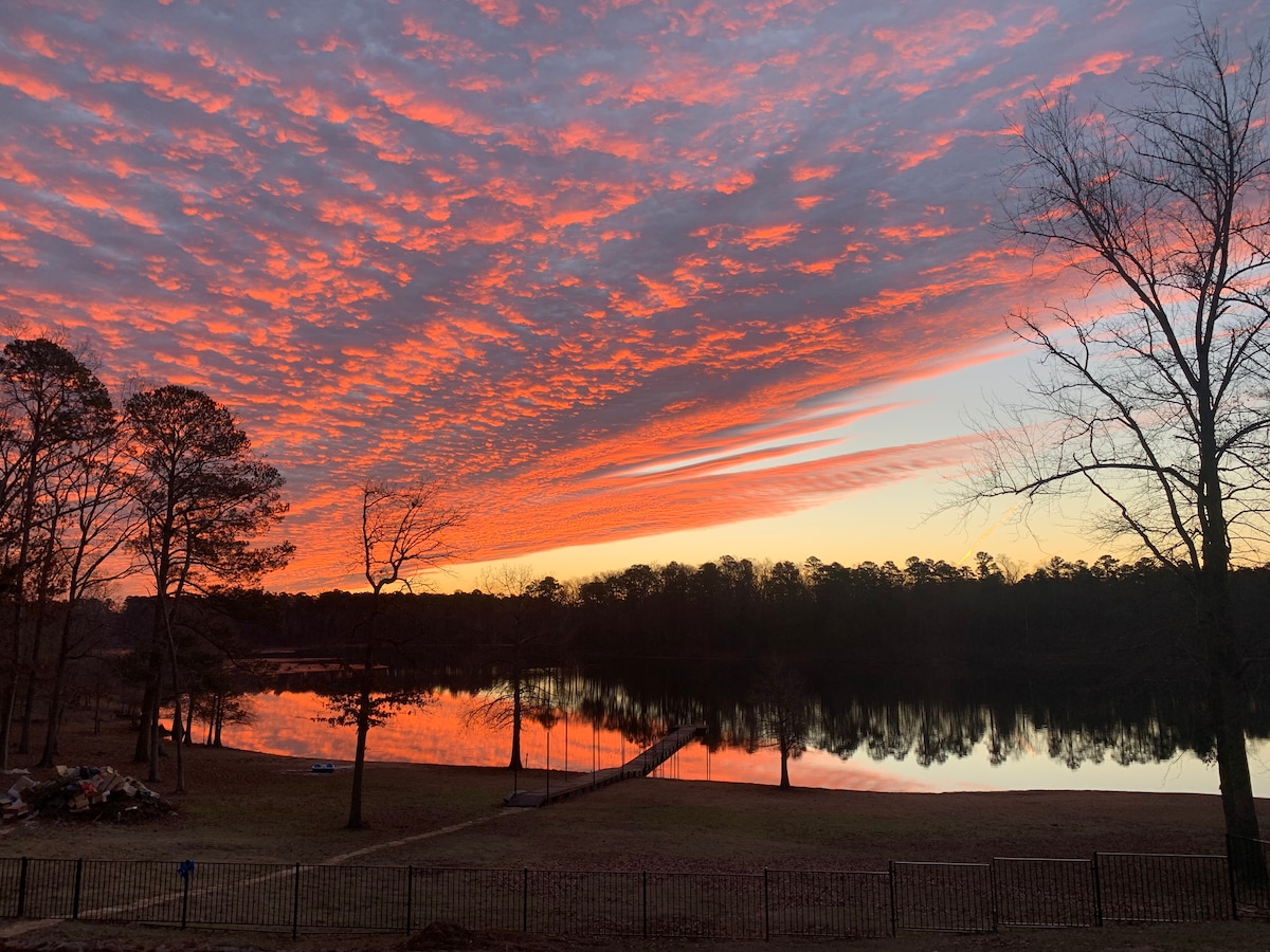 A vibrant sunset reflects across the calm surface of the lake, with a colorful array of oranges and pinks filling the sky. Silhouetted trees frame the scene, and a fishing pier can be seen extending into the water, providing a serene backdrop.