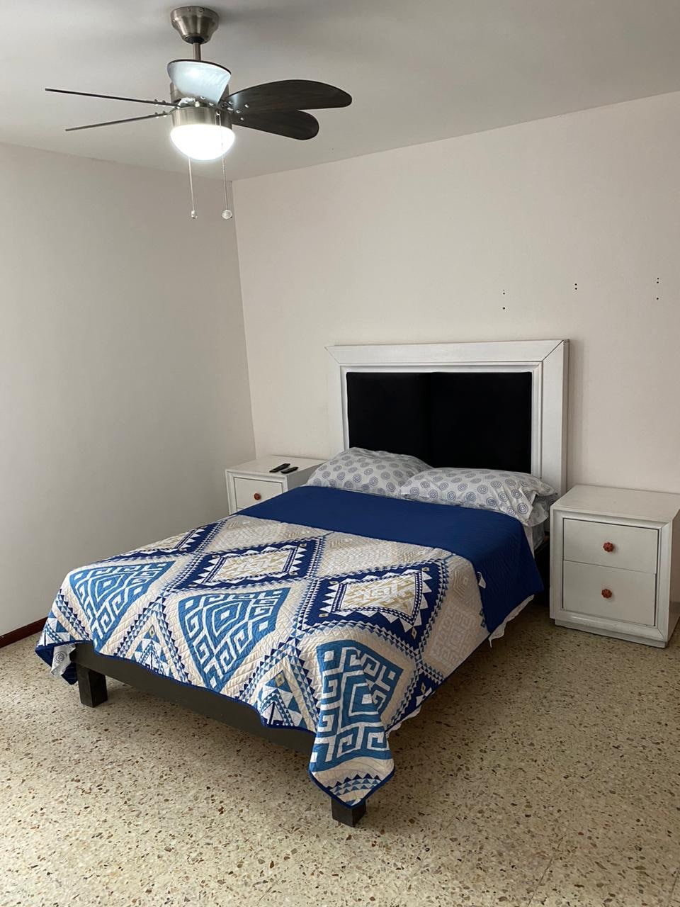 A cozy sleeping area features a spacious bed adorned with a blue and white patterned blanket. Two bedside tables flank the bed, each equipped with a small drawer. A ceiling fan is mounted above, providing an airy feel to the room.