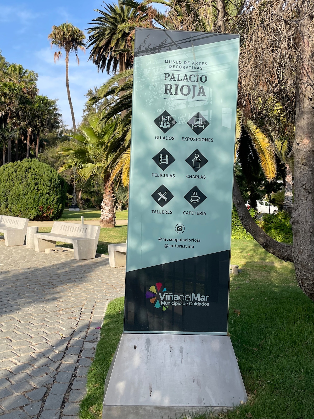 A large informational sign is set in a park-like environment, displaying details about the Museo de Artes Decorativas Palacio Rioja. The sign includes symbols indicating services such as guided tours, exhibitions, movies, and workshops, surrounded by greenery and palm trees.