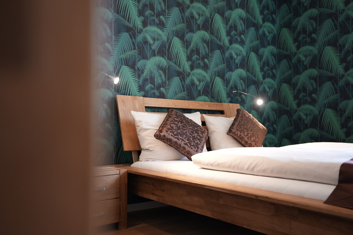 A cozy bedroom features a wooden bed frame with soft bedding and decorative pillows. The wall is adorned with a leafy green wallpaper, while bedside lamps provide subtle illumination. A wooden nightstand accompanies the bed, contributing to the room's warm atmosphere.