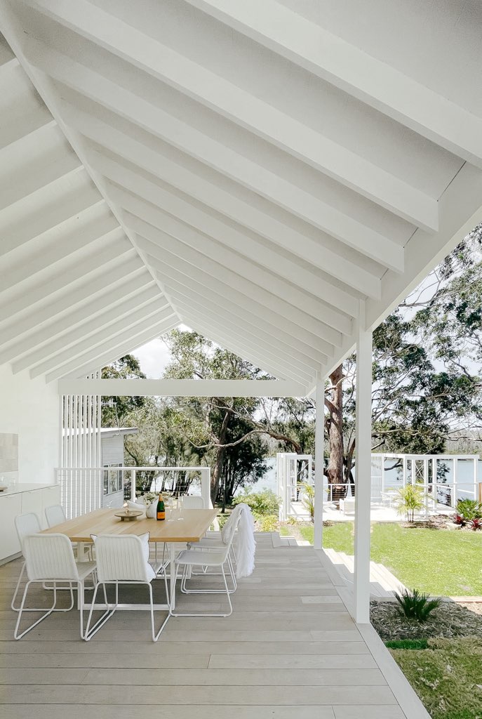An outdoor dining area features a long wooden table surrounded by white metal chairs. The space is sheltered beneath a sloped roof with exposed beams. Lush greenery is visible in the background, along with a glimpse of the water's edge.