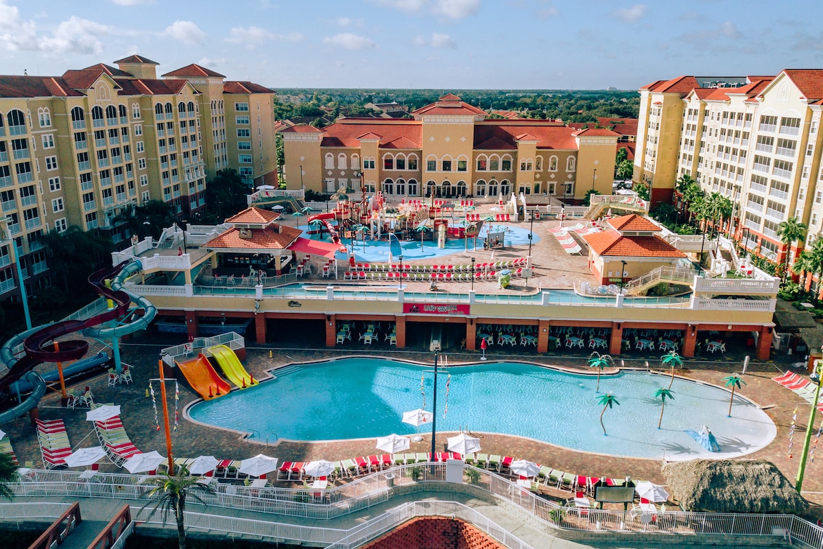 The expansive outdoor pool area features a large, zero-entry pool and colorful water slides. Lounge chairs line the poolside, with shaded areas and palm trees enhancing the inviting setting. The resort buildings stand in the background, showcasing a blend of vibrant colors beneath a clear blue sky.