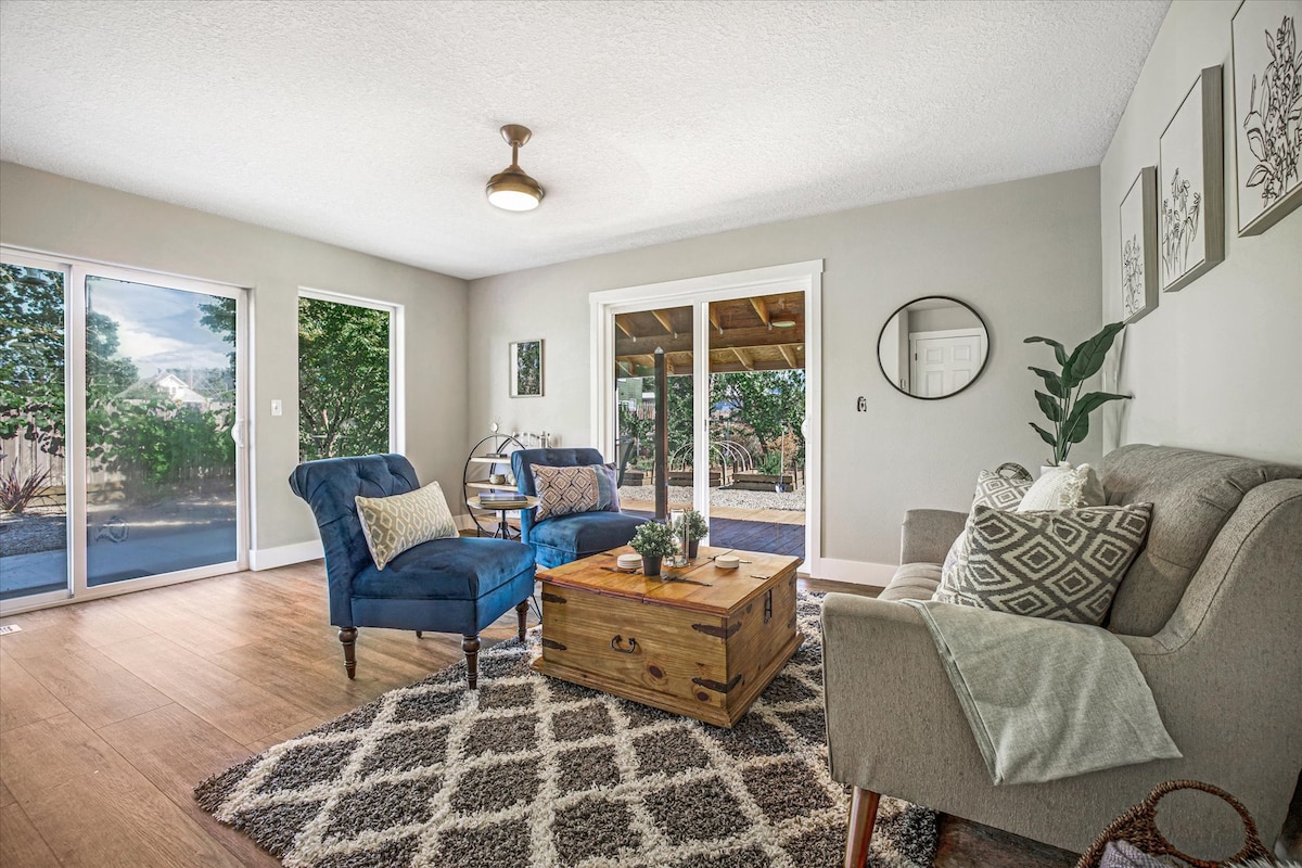 A cozy living area is presented with two blue armchairs and a light grey couch, complemented by a wooden coffee table. Natural light fills the room through large glass doors that open to the outdoor area, enhancing the welcoming atmosphere.