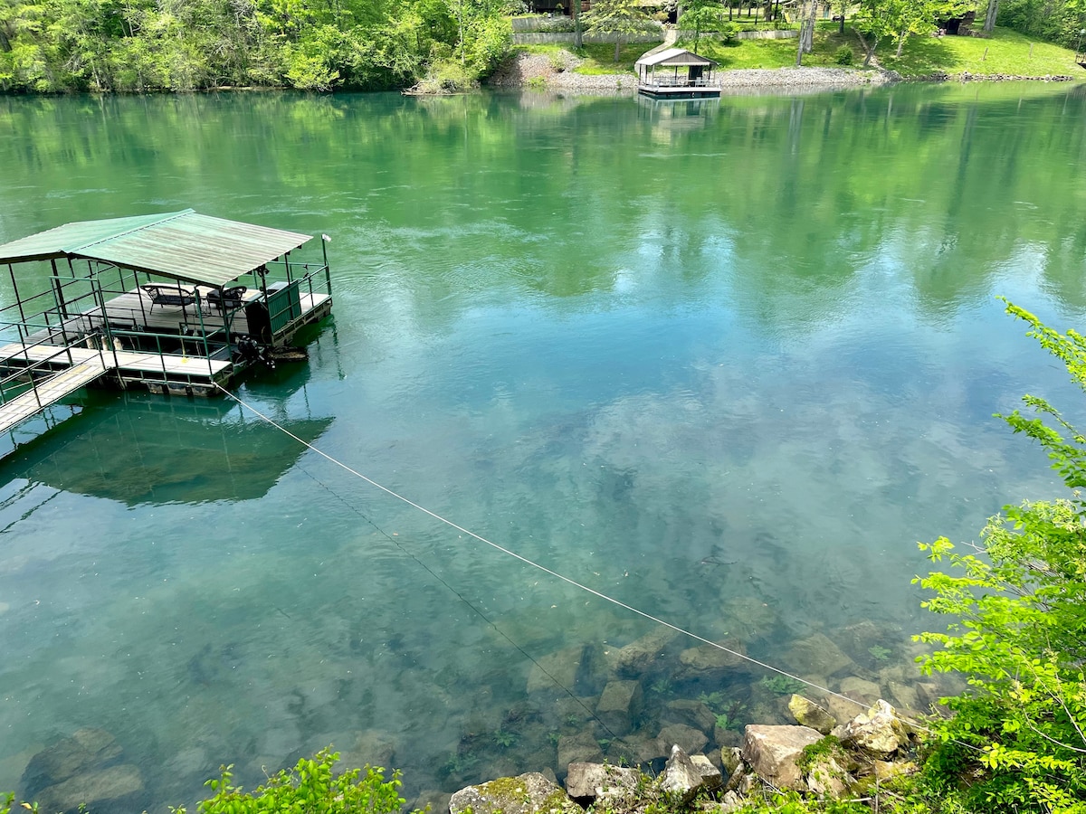 A serene view of the Little Red River is presented, featuring calm, clear waters reflecting surrounding greenery. A private boat dock is visible close to the riverbank, offering easy access to water activities, while a wooden structure stands in the distance, enhancing the natural setting.