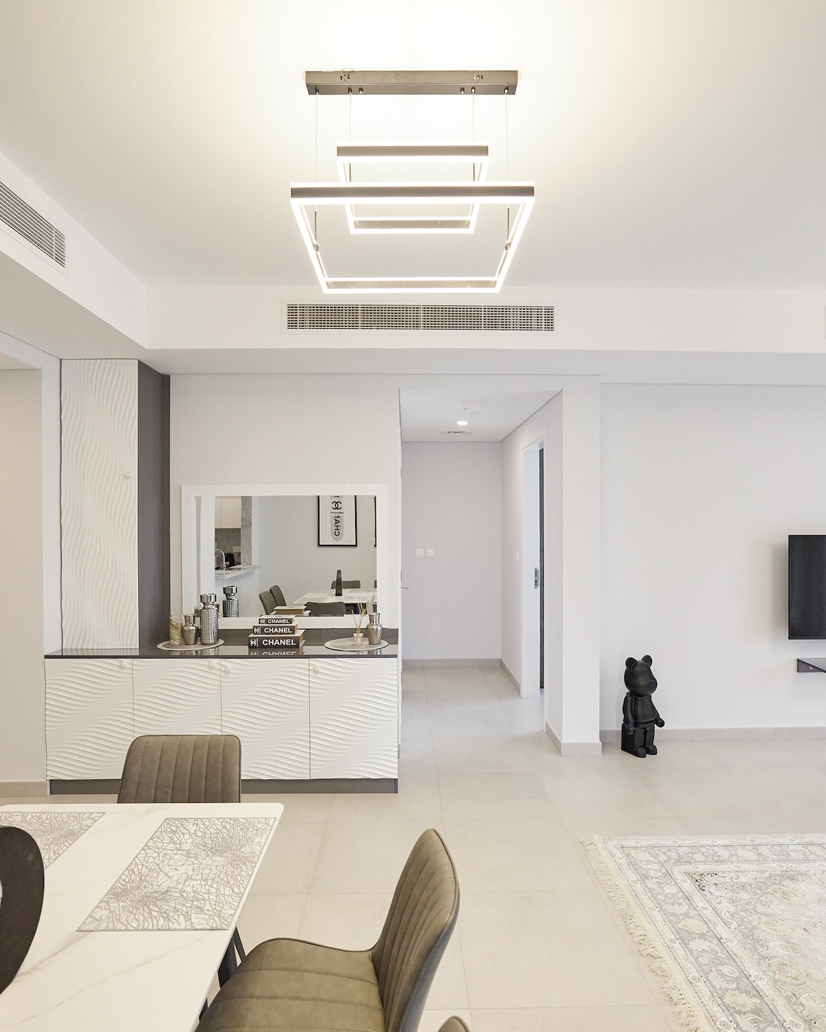 A modern dining area features a minimalist table accompanied by stylish chairs, with a striking geometric light fixture above. A neatly arranged sideboard holds various items, reflecting a clean and spacious design. To the side, a sleek television is mounted on the wall.