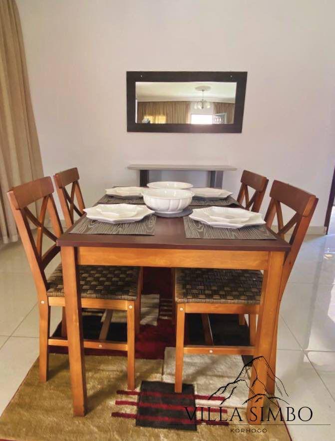 A dining area is presented with a wooden table surrounded by four chairs. A bowl sits at the center of the table, complemented by placemats. The wall features a large mirror reflecting the space, and natural light flows through nearby curtains.