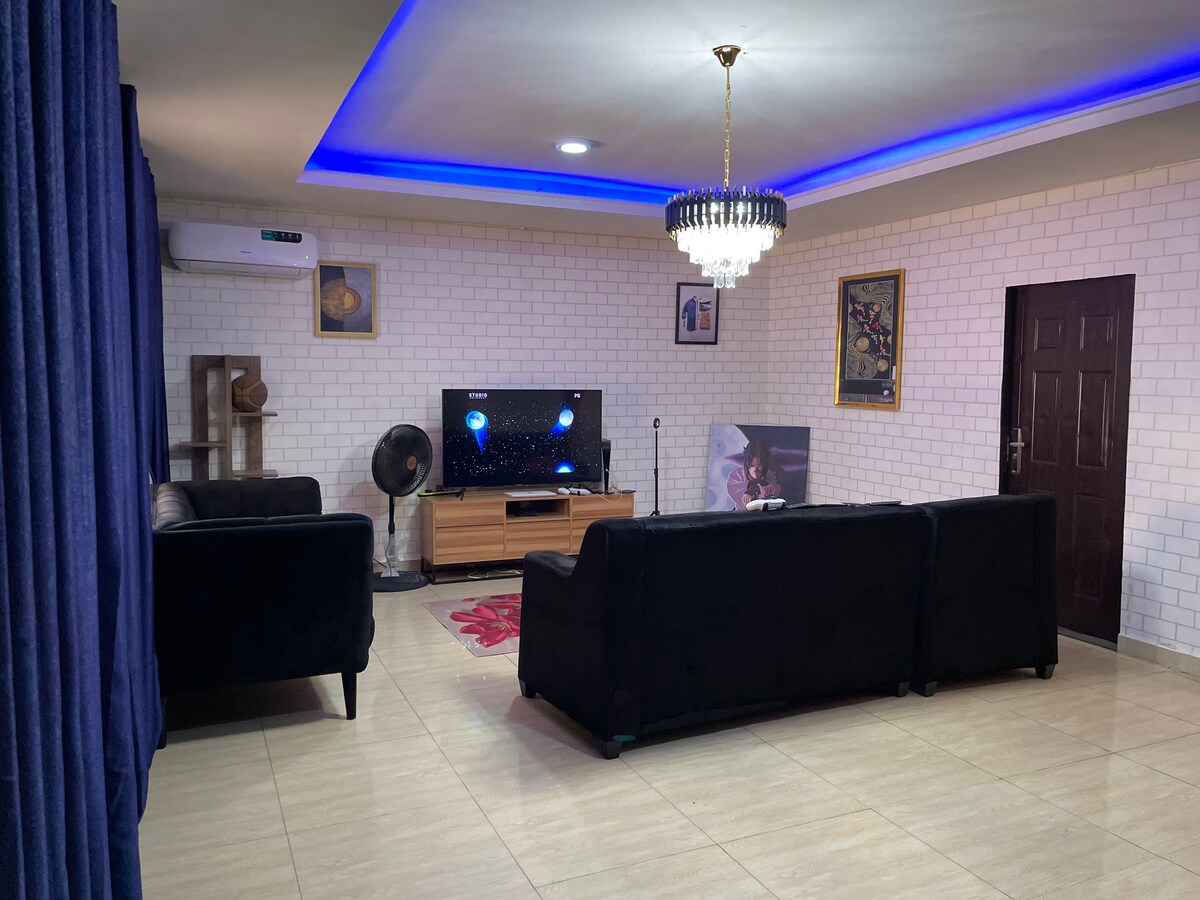 A spacious living room features dark, plush sofas positioned around a wooden media unit with a large television. A decorative chandelier hangs from the ceiling, while a fan sits beside the seating area. The walls are adorned with framed artwork, and a soft area rug adds comfort.