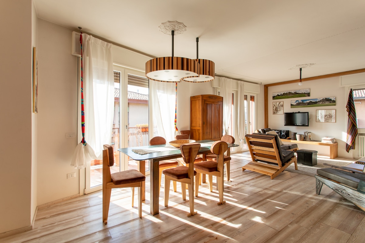 A spacious dining area is furnished with a wooden table surrounded by matching chairs. Two modern light fixtures hang above, providing illumination. Large windows allow natural light to filter in, with a cozy seating area visible in the background, featuring a flat-screen TV and decorative wall art.