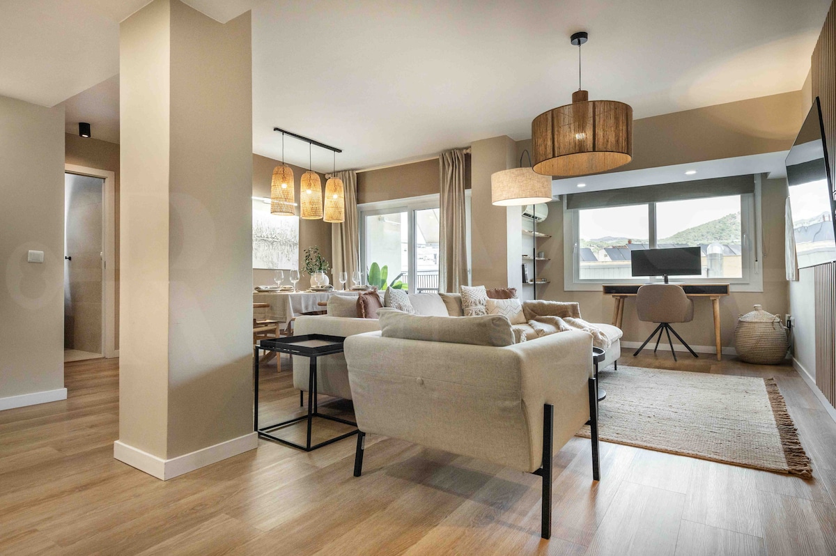 A spacious living area is presented with a light neutral color scheme. Comfortable seating options include a sofa and armchair, complemented by a dining table for six. Large windows allow natural light to fill the space, with a stylish desk and smart TV visible.