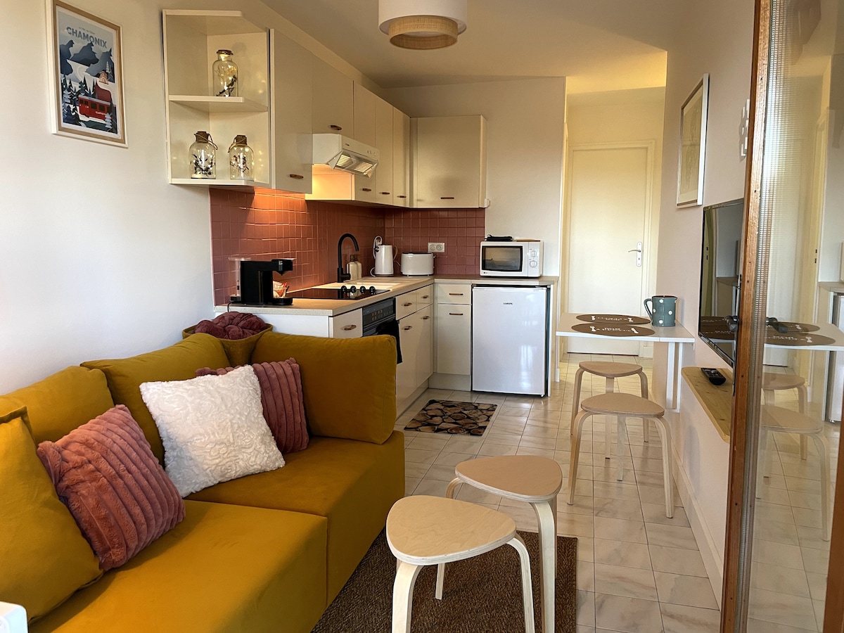 An open-plan living area features a cozy yellow sofa with decorative pillows, complemented by a light wooden coffee table set on a woven rug. A well-equipped kitchen area showcases modern appliances and cabinetry, with a dining table for two positioned nearby.