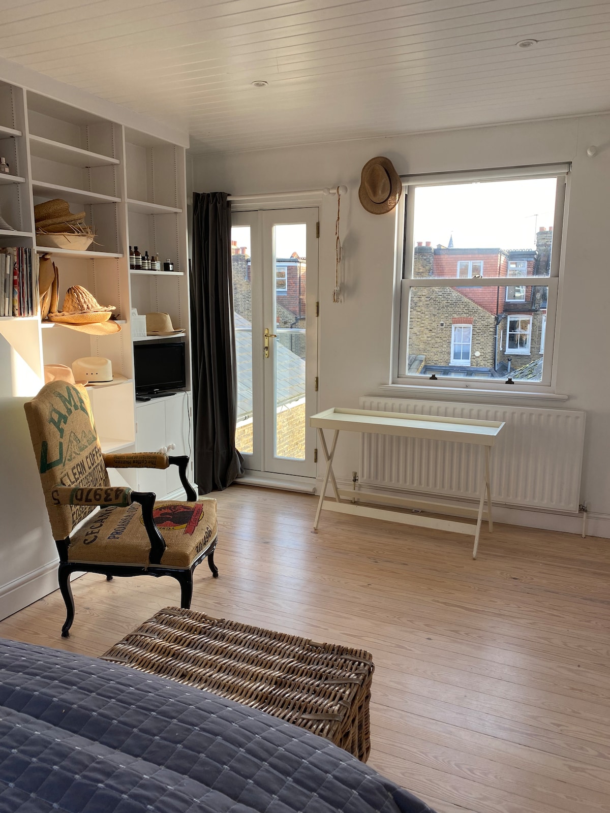 A stylishly decorated room features a chair with an artistic pattern and a compact table positioned near a window. Natural light streams in, illuminating the wooden floor. Shelving with decorative items and personal care products is visible on the opposing wall.