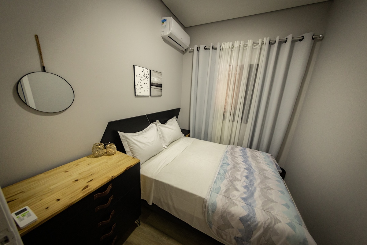 The cozy bedroom features a spacious bed with white linens, complemented by a decorative patterned throw. A wooden dresser stands beside the bed, while soft curtains frame the window, allowing natural light to enter the room. A round mirror adds depth to the space.