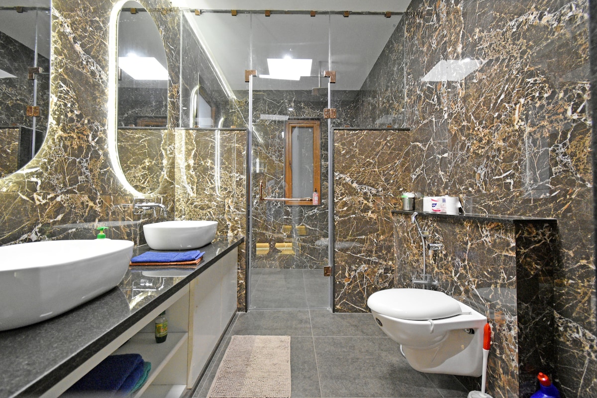 A spacious bathroom features elegant marble walls and flooring, complemented by two modern sinks situated on a dark countertop. Glass partitions separate the shower area, with a large rain showerhead above. A discreet toilet is positioned against the wall, ensuring a sleek design.
