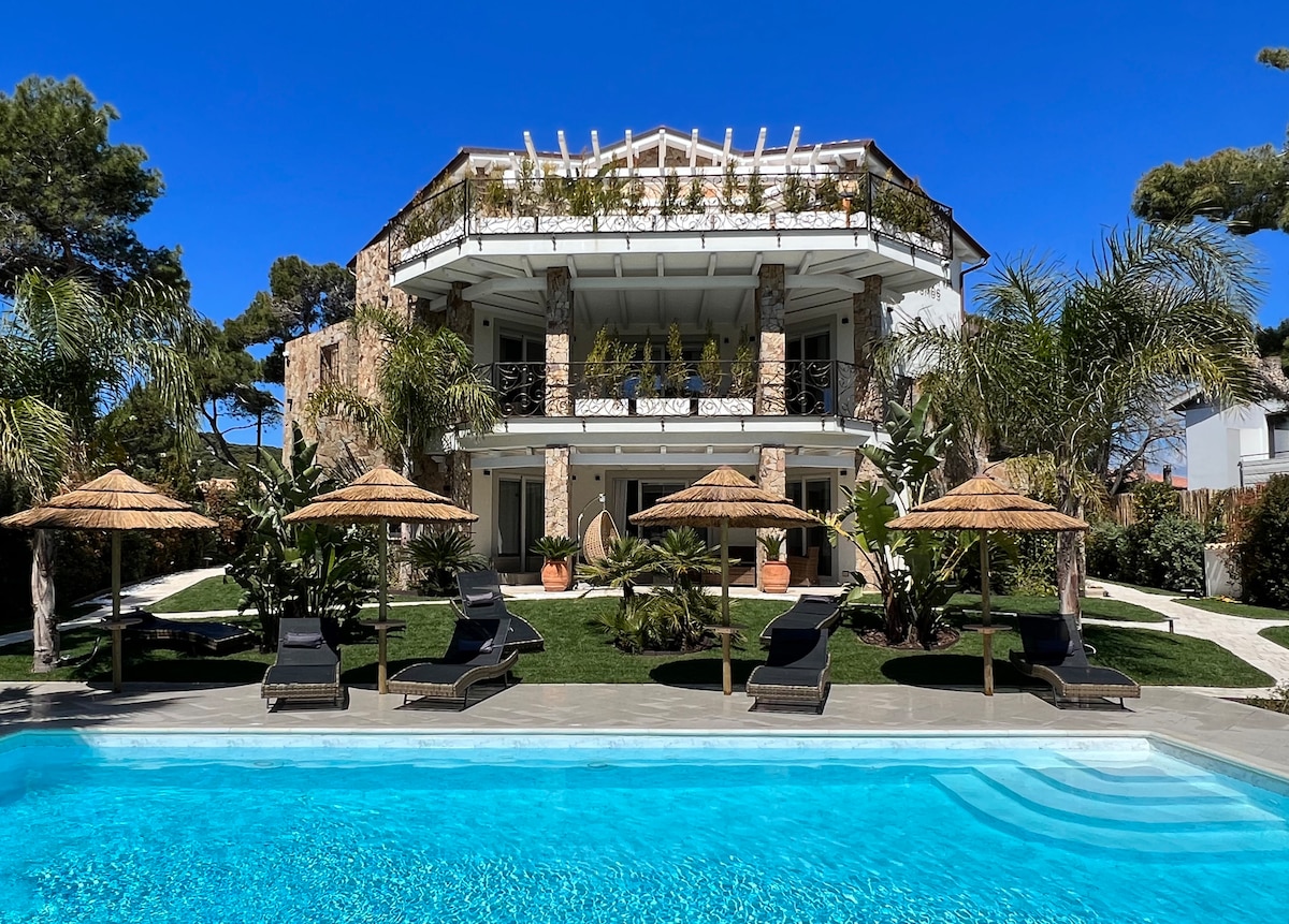 The villa showcases a modern architectural design with stone accents and multiple levels. A clear blue pool is in the foreground, bordered by sun loungers under thatched umbrellas. Lush greenery and well-maintained gardens surround the property, offering a serene outdoor space.