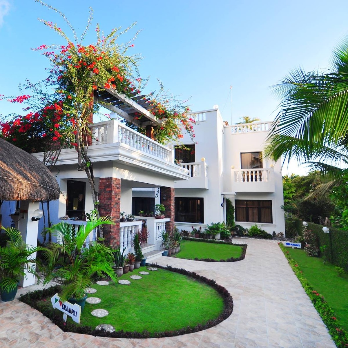 The exterior of a modern hotel features a well-maintained garden with lush greenery and flowering plants. A spacious terrace with a railing is visible, and the building is accented with stone and brick details. Pathways are lined with decorative stones leading to the entrance.