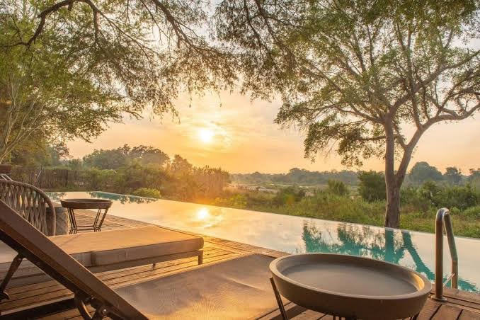 A tranquil outdoor space features lounge chairs positioned around a pool, with a wooden deck extending towards a landscape of greenery. The setting sun casts a warm glow over the horizon, illuminating the natural surroundings and creating a peaceful ambiance.