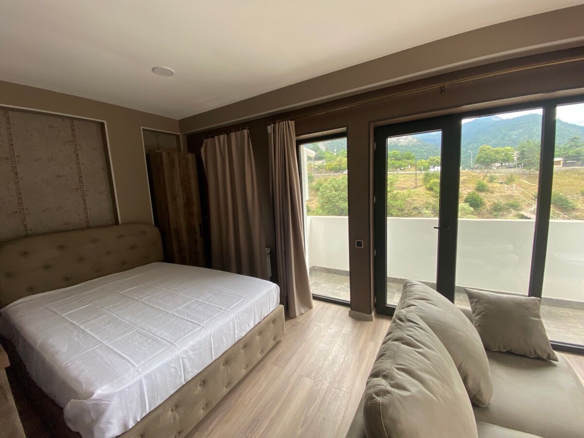 A serene bedroom is depicted, featuring a spacious bed with a neutral-colored headboard. Large glass doors open to a balcony, allowing natural light to fill the room. Nearby, a comfortable couch adds a cozy touch, with local greenery visible outside.