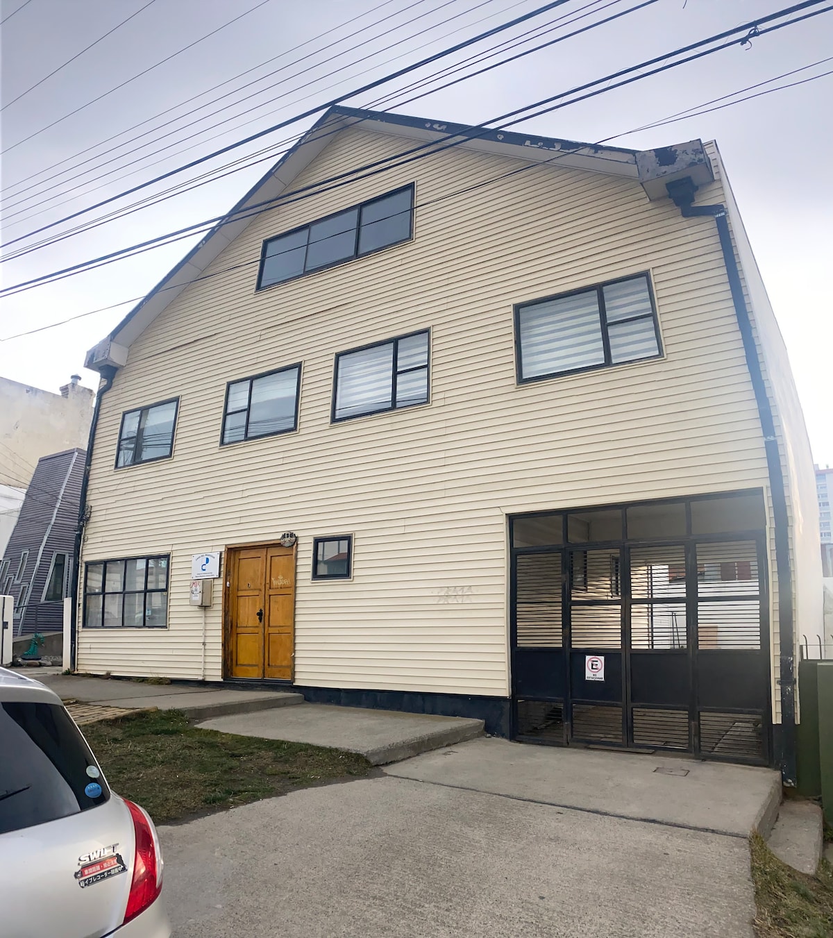 The exterior of a two-story building is displayed, featuring a light-colored facade with large, rectangular windows. A wooden door is positioned centrally at the front, while a covered entryway is visible to the right. A small garden area is located in front of the building.