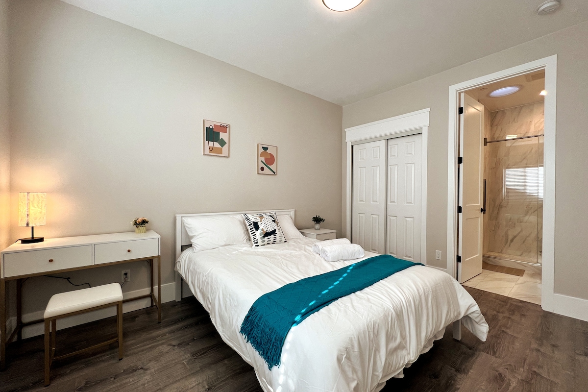A well-appointed bedroom features a spacious bed with crisp white linens and a decorative throw. A small desk and chair are situated beside the bed. The en suite bathroom is visible through a partially opened door, showcasing modern fixtures and marble accents.