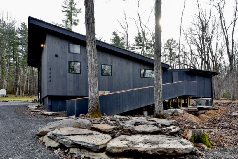 A contemporary home with a striking black exterior is set among tall trees. A ramp leads to the entrance, surrounded by large flat stones on the ground. The structure features multiple windows, allowing natural light to enter while blending with the natural surroundings.