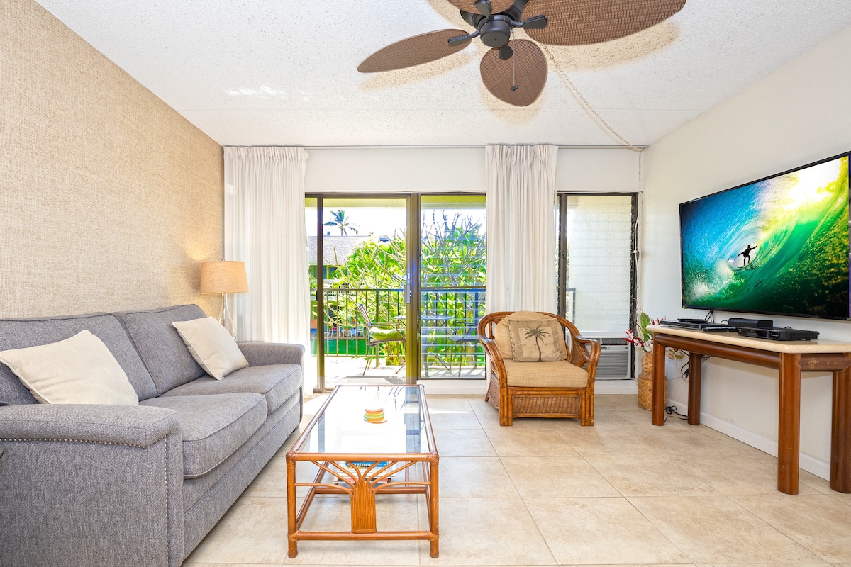 The living room features a gray sofa, a rattan armchair, and a glass coffee table. Natural light filters in through sliding glass doors, leading to a private balcony. A flat-screen TV is mounted on the wall, with a ceiling fan above providing added comfort.