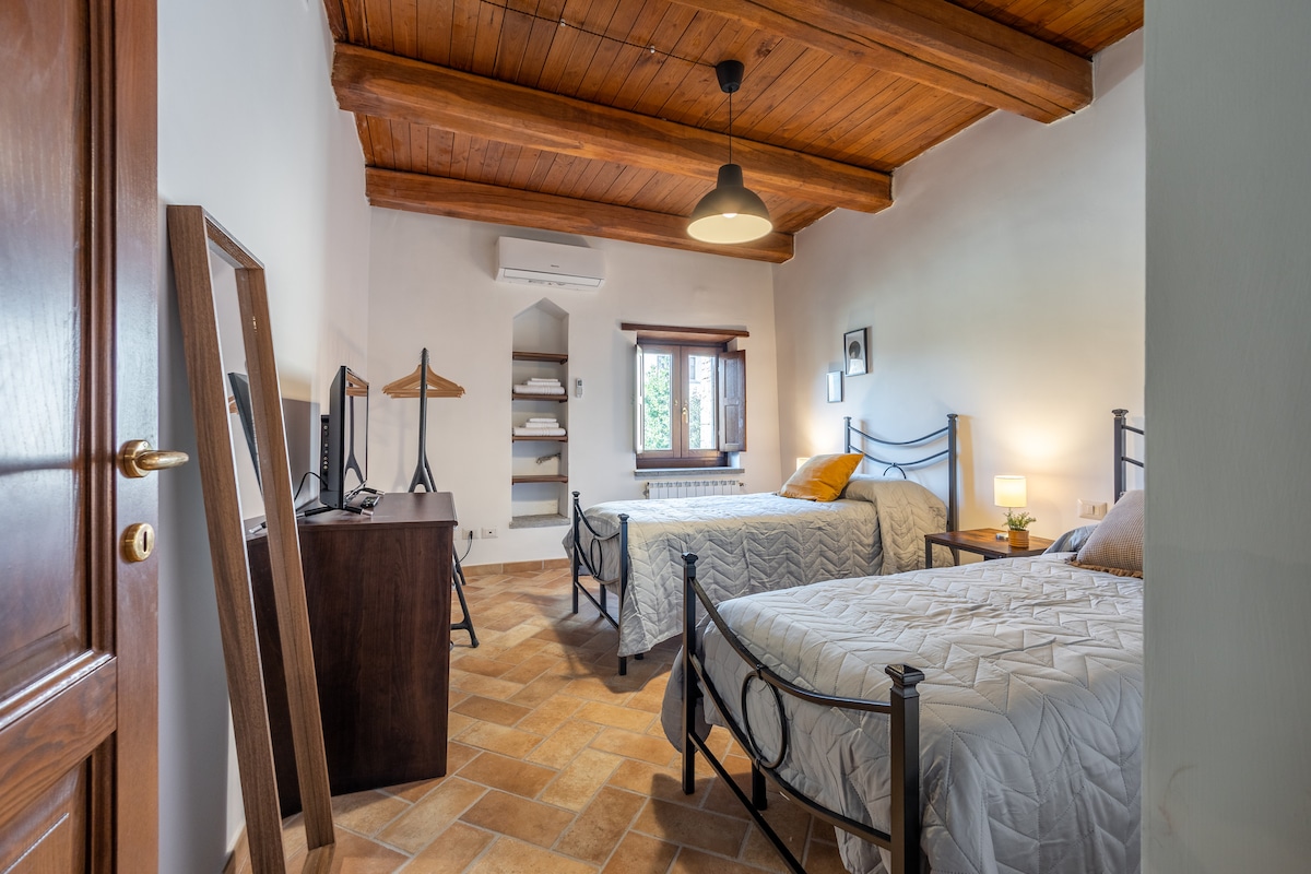 A well-lit bedroom featuring two single beds with light bedding, framed by rustic wooden beams on the ceiling. A dresser and mirror are positioned nearby, while a small desk with a television is in the corner. Natural light filters through the window with wooden shutters.