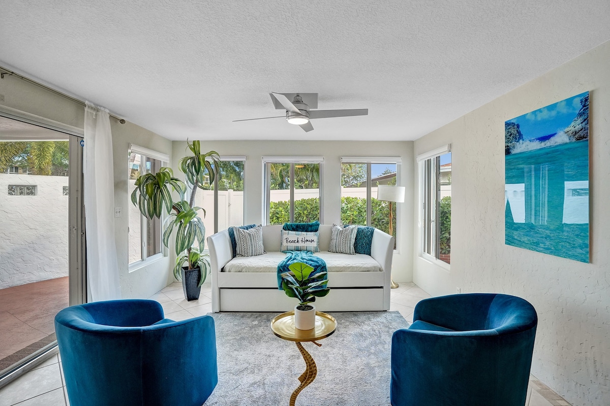 A bright living area features a day bed adorned with a decorative pillow, surrounded by two blue chairs and a round gold-accent table. Large windows allow natural light to fill the space, while greenery is present in the corner, adding a touch of nature to the room.