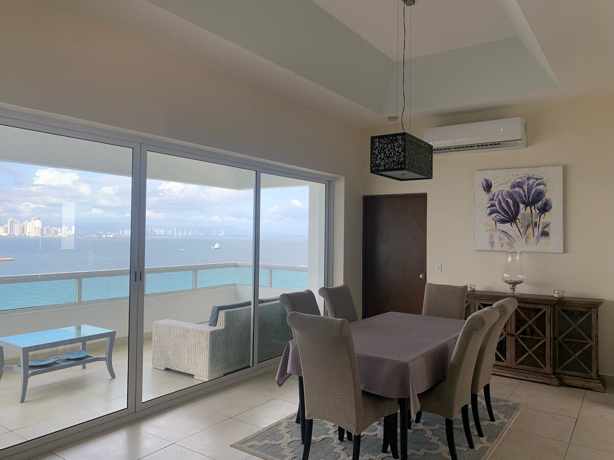 A dining area is seen with a large table surrounded by upholstered chairs. A soft rug complements the space. The backdrop presents a panoramic view of the ocean and Panama City through floor-to-ceiling sliding glass doors, framed by gentle natural light.