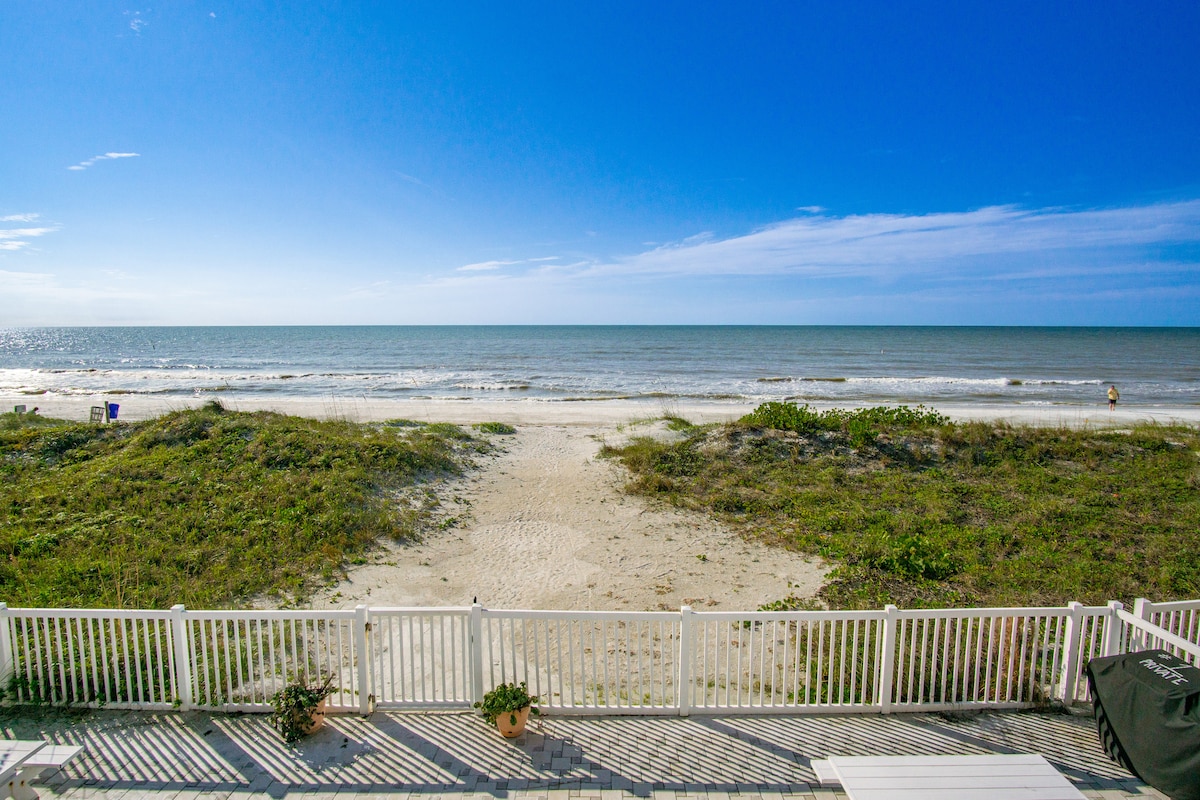 An expansive view of the ocean stretches out under a clear blue sky. Soft sandy beach is visible in the foreground, with gentle waves lapping the shore. A white fence outlined with greenery frames the scene, creating a serene coastal environment.