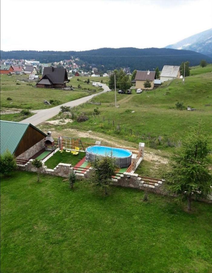 A spacious outdoor area is featured, with a round above-ground pool surrounded by a grassy lawn. Colorful play equipment is present, along with nearby trees and a scenic view of the mountains and distant houses. A gravel path leads through the serene landscape.