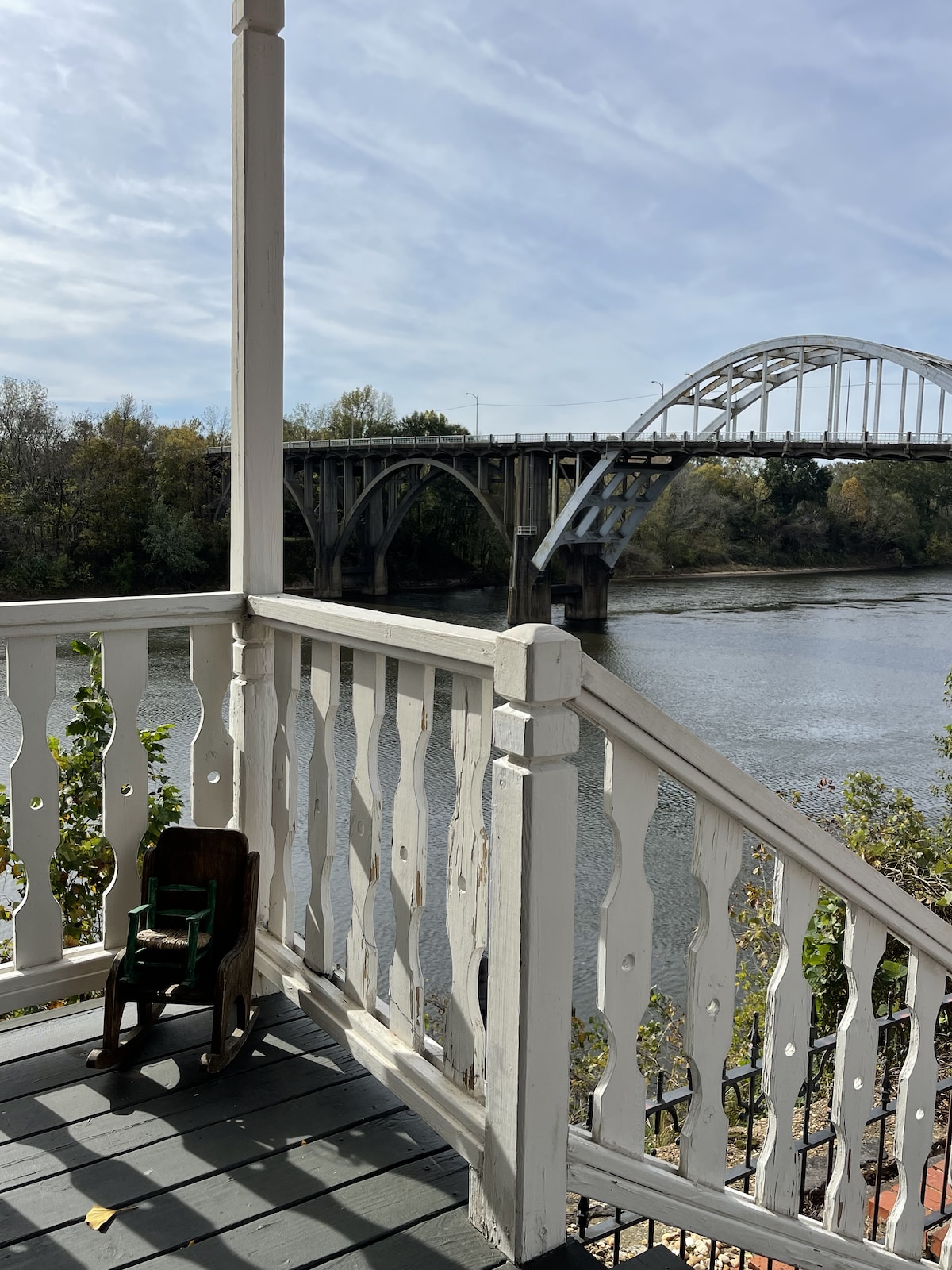 A private balcony offers scenic river views and a direct sightline to the iconic Edmund Pettus Bridge. The wooden railing features decorative cutouts, and a small, vintage rocking chair adds a charming touch. Lush trees line the riverbanks, and the water reflects the sky.