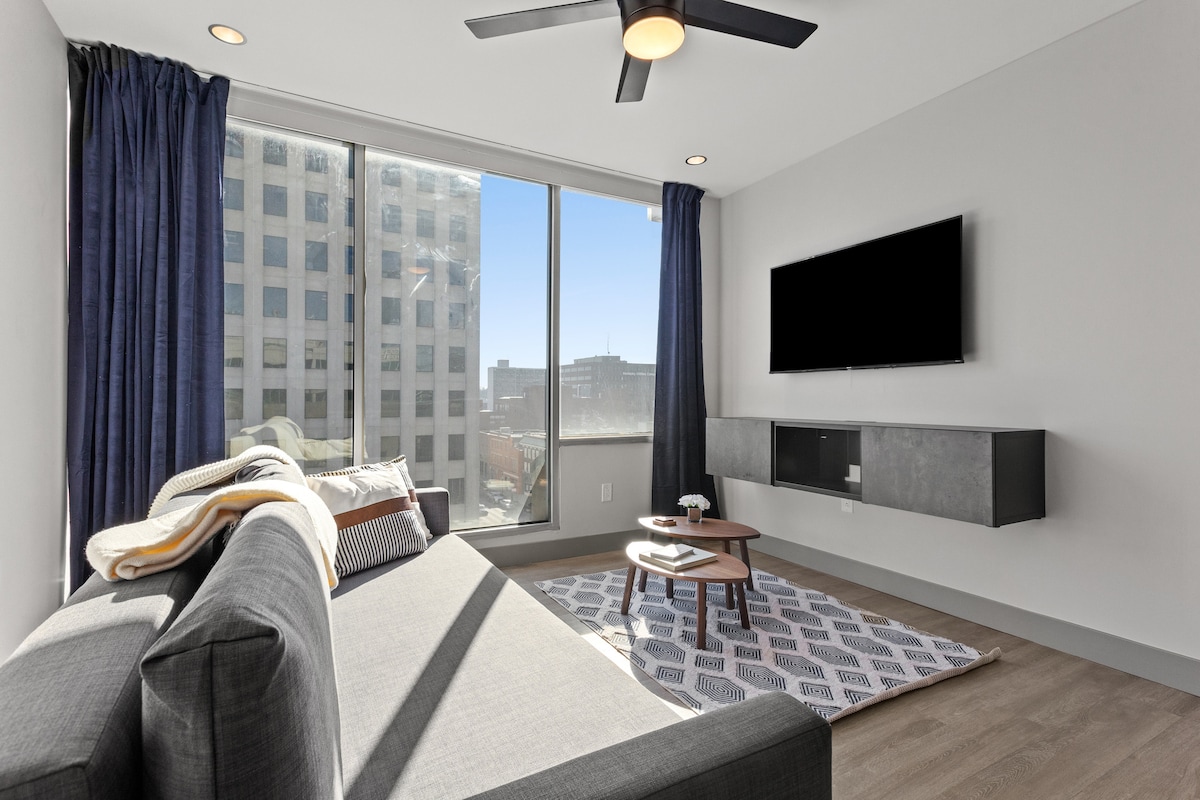 A modern living room features a comfortable gray sofa positioned near large windows that invite ample natural light. A small coffee table is centered on a patterned rug, while a flat-screen television is mounted on the wall. Soft blue curtains frame the windows.