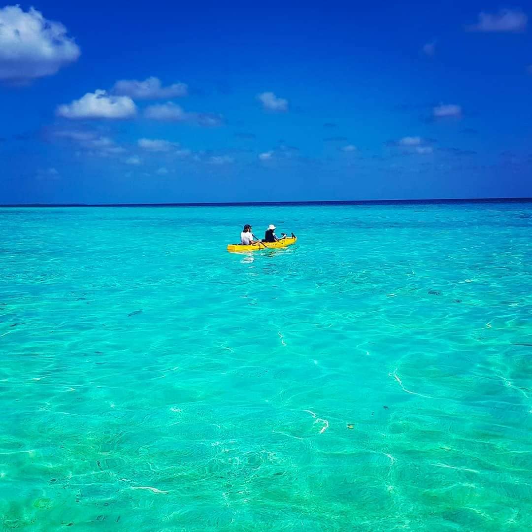 A yellow kayak occupies the clear turquoise water, with two individuals seated inside. The calm sea reflects the bright blue sky, dotted with a few fluffy clouds. The serene environment creates an inviting scene for water activities.