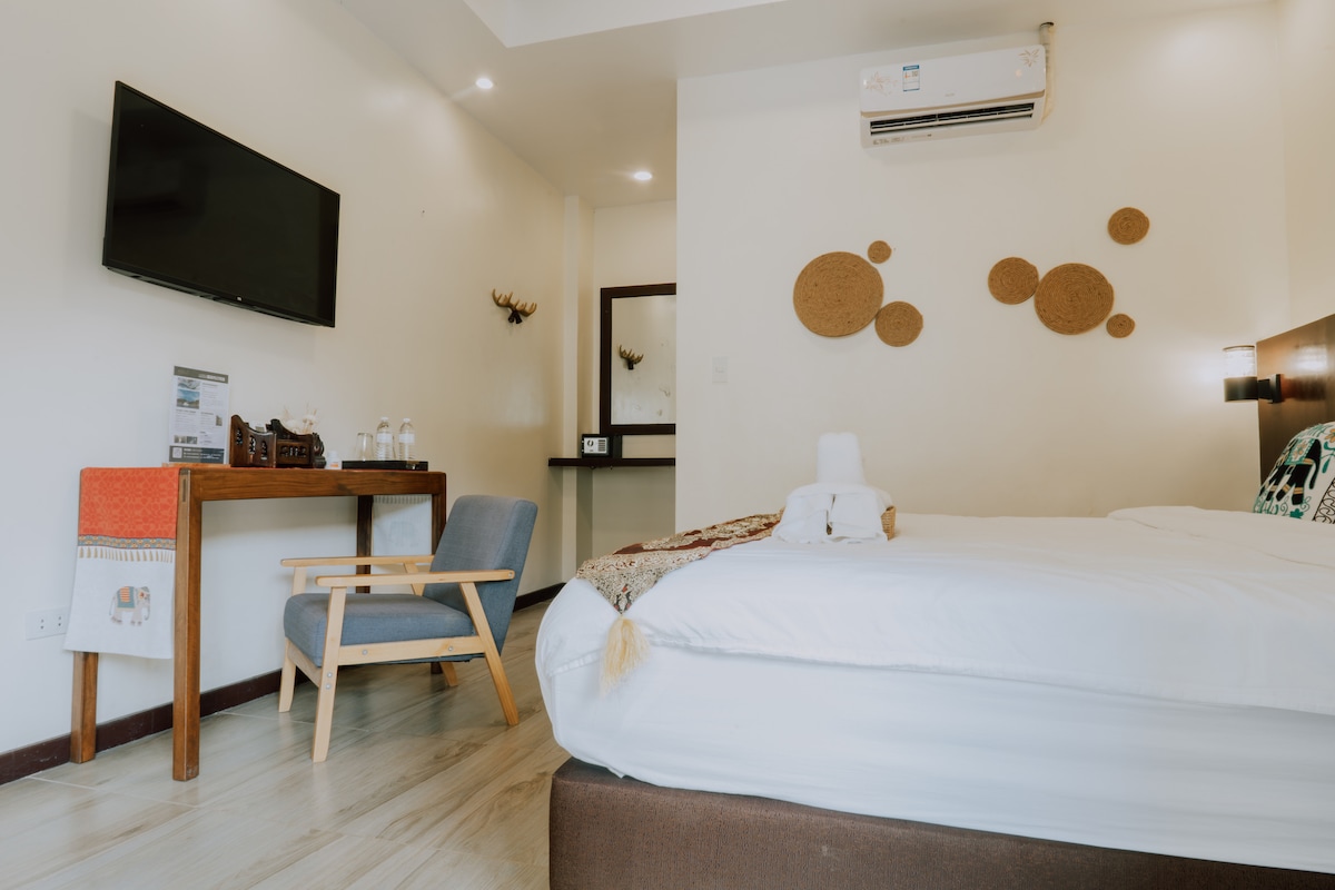 A modern guest room is presented with a spacious bed and a flat-screen television mounted on the wall. A small wooden desk with a chair is positioned beside the bed, while decorative wall accents enhance the ambiance. Soft lighting illuminates the room.