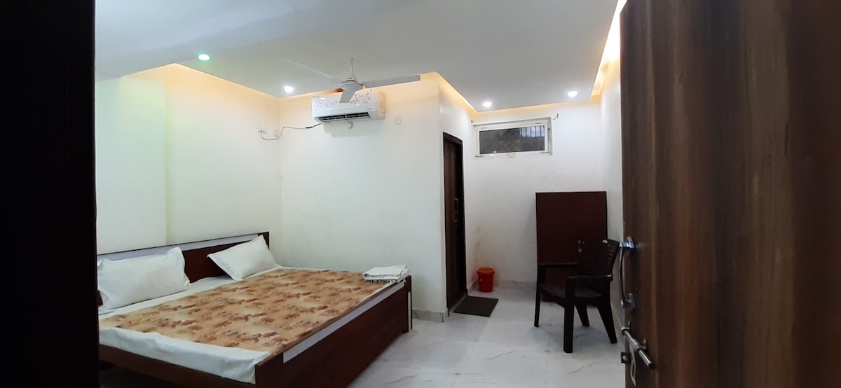 A spacious bedroom features a large bed with neutral bedding and pillows, accompanied by a wooden chair and a table. Soft lighting is provided by ceiling fixtures, enhancing the serene atmosphere. A fan is installed on the ceiling, and a door leads to an additional area.