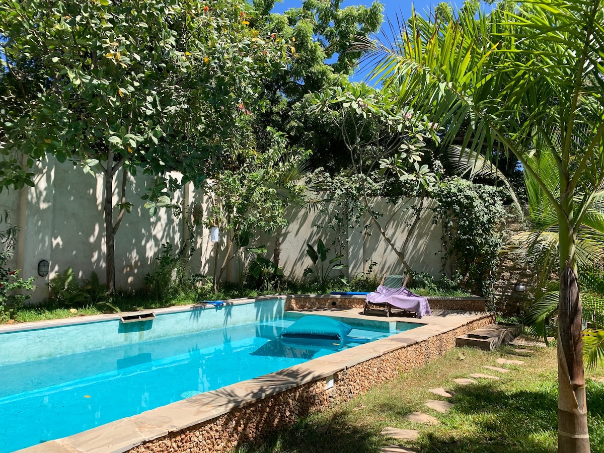 A serene outdoor space is featured with a crystal-clear swimming pool surrounded by lush greenery. Smooth stone pathways lead to shaded areas, while vibrant palm trees and flowering plants add to the peaceful ambiance of the setting.