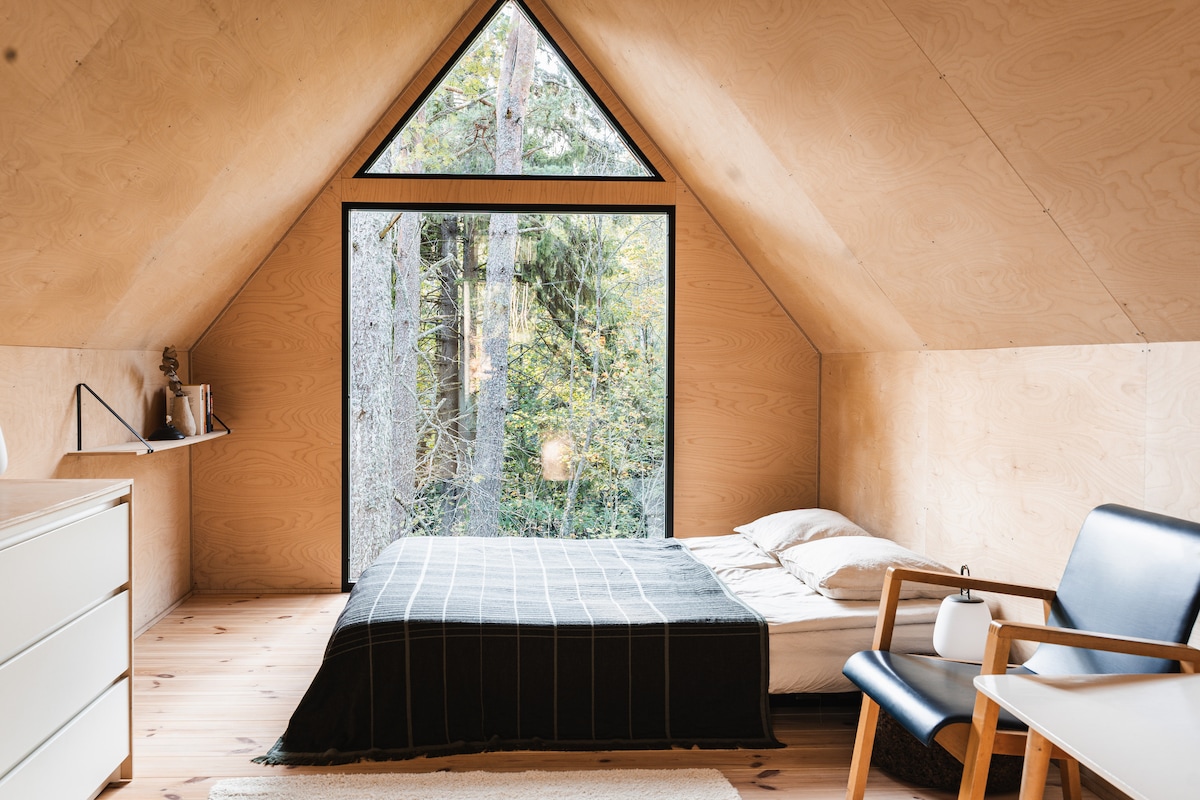 A minimalist bedroom features a spacious bed adorned with a dark blanket, positioned near a large triangular window that overlooks the surrounding forest. Light wooden walls create a warm ambiance, complemented by simple furnishings, including a chair and dresser.