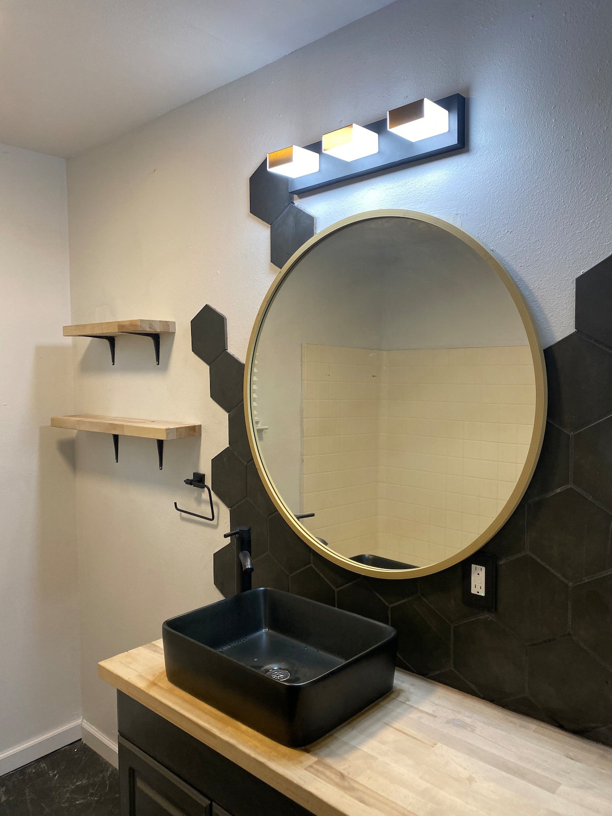 A modern bathroom is showcased, featuring a round mirror with a gold trim above a sleek black basin. Hexagonal tile accents the wall, complemented by two wooden shelves. Soft lighting is provided by a three-bulb fixture mounted above the mirror.