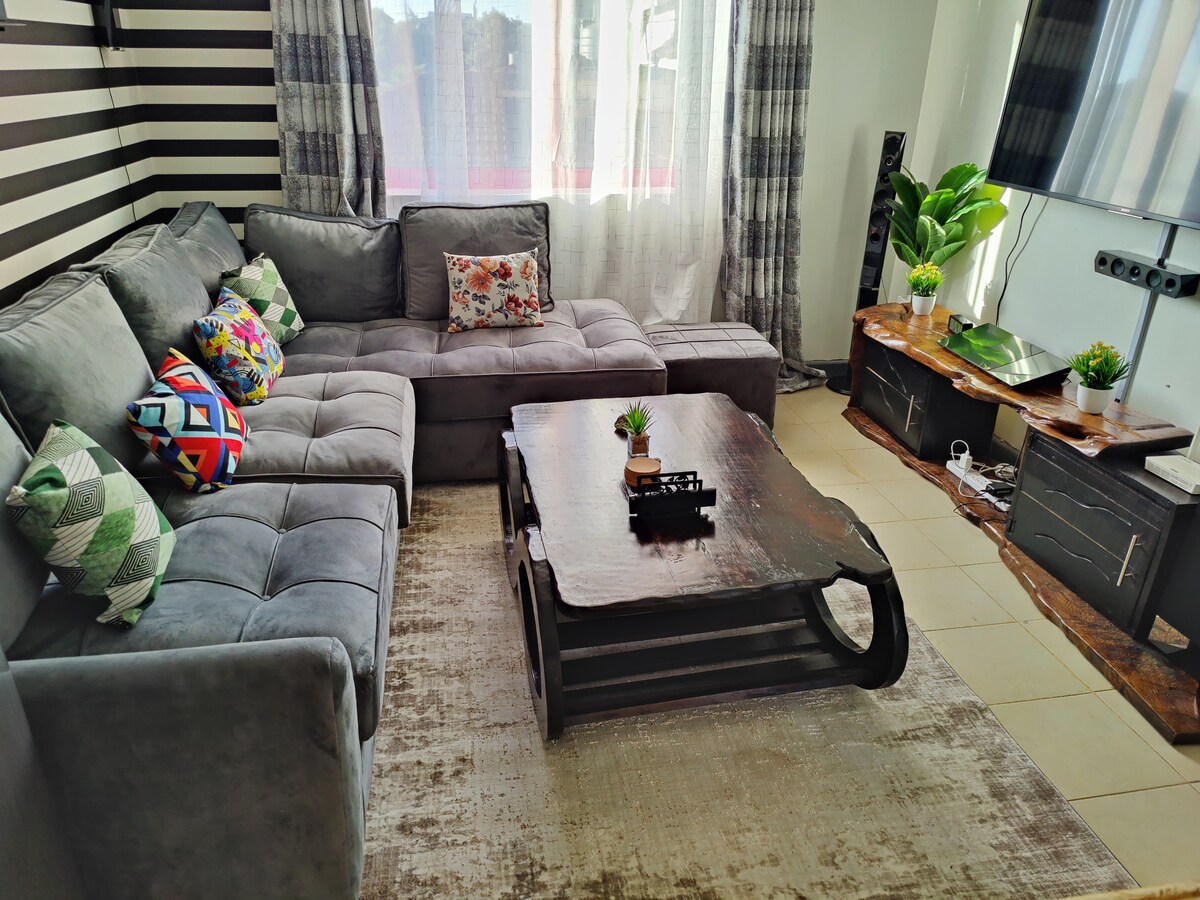 A spacious living area features a large sectional gray sofa adorned with colorful accent pillows. A wooden coffee table with a decorative plant sits in the center, complemented by a neutral area rug. Sunlight streams through large windows, enhancing the relaxed ambiance.