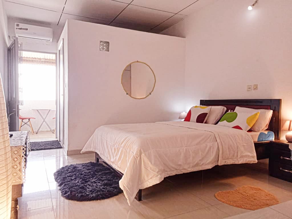 A well-lit sleeping area features a spacious bed with colorful accent pillows. A circular mirror is mounted on the wall, and soft rugs lie on the tiled floor. Natural light enters through a nearby window, enhancing the minimalist decor.