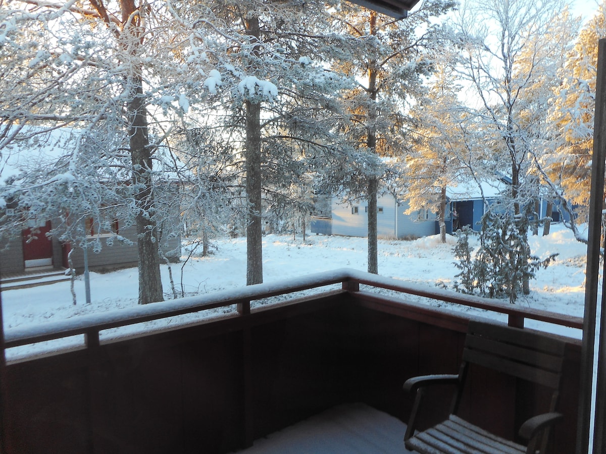 A snow-covered balcony offers a view of frost-laden trees and a serene winter landscape. A wooden chair is positioned on the balcony, inviting quiet moments in nature. Nearby structures are visible, surrounded by the tranquil beauty of freshly fallen snow.