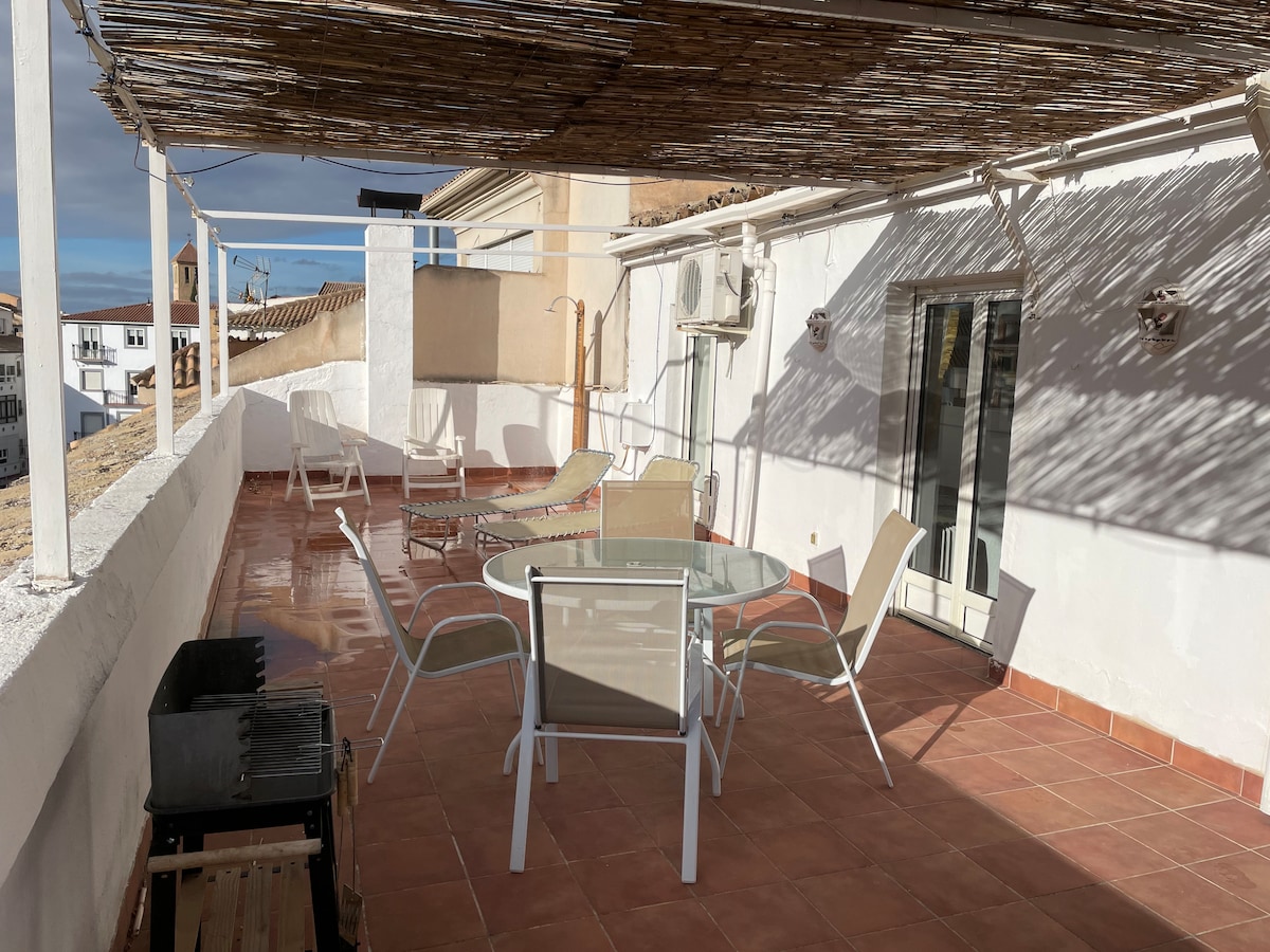 A spacious terrace is featured with a circular glass table surrounded by white chairs. Two loungers are positioned for relaxation. A barbecue grill is visible to one side, and light patterns are created on the floor by a thatched awning overhead.