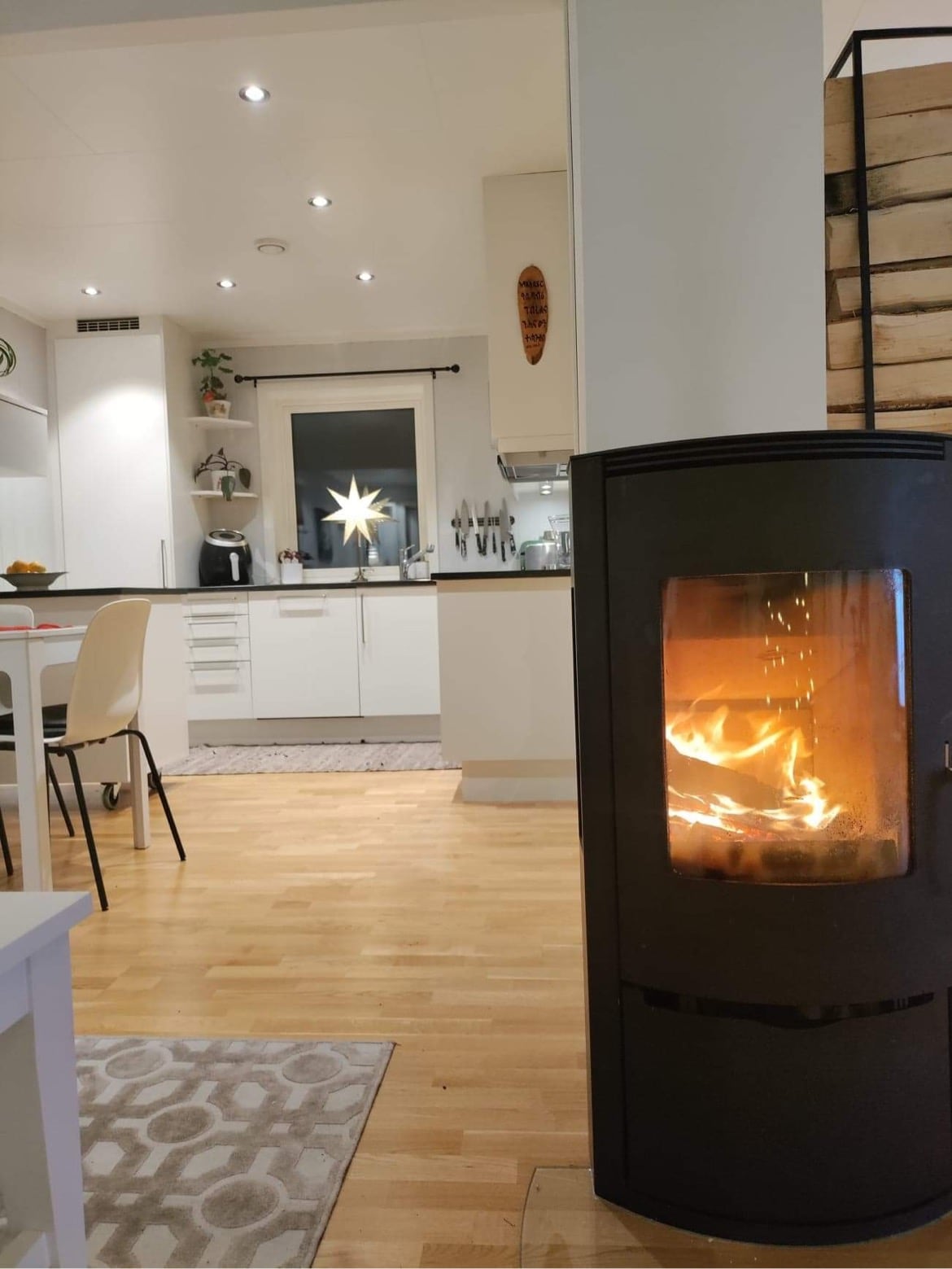 A modern kitchen is visible in the background, featuring sleek cabinetry and a bright atmosphere. A wood-burning stove stands prominently in the foreground, with flames flickering gently. A dining table is set nearby, creating an inviting and warm space.
